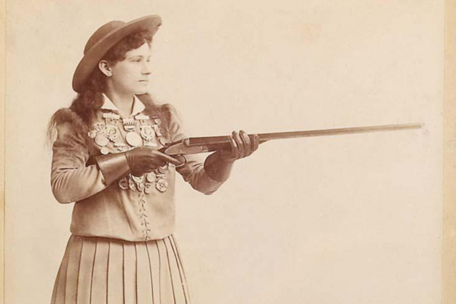 Sepia-toned portrait of a woman in early Western attire, wearing a pleated skirt, gloves, and a wide-brimmed hat, confidently holding a rifle. Her jacket is adorned with numerous medals, suggesting she is a decorated markswoman, likely from the late 19th or early 20th century.
