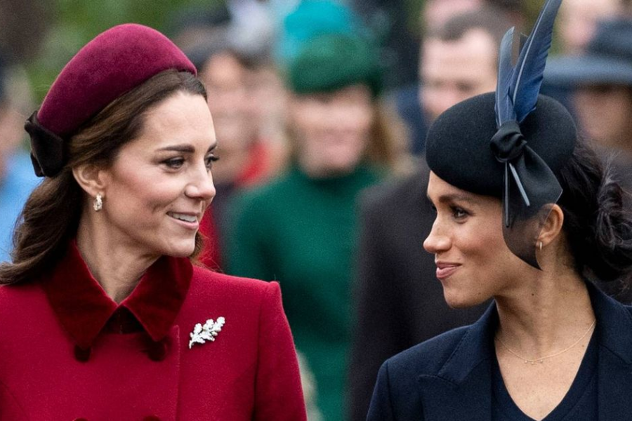 Two women dressed in elegant winter coats and matching hats share a smile during a public event. The woman on the left wears a deep red outfit with a brooch and pearl earrings, while the woman on the right wears a navy ensemble with a feathered hat.