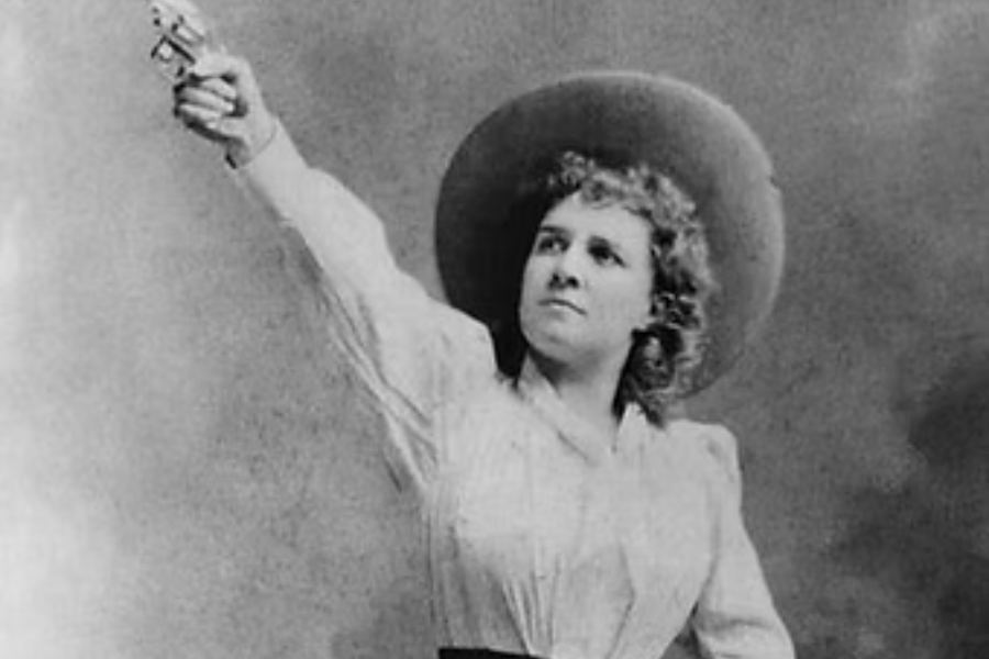 Black and white portrait of a confident woman in a wide-brimmed hat raising a revolver high in her right hand. She wears a blouse with puffed sleeves and gazes upward with determination, striking a bold and dramatic pose.