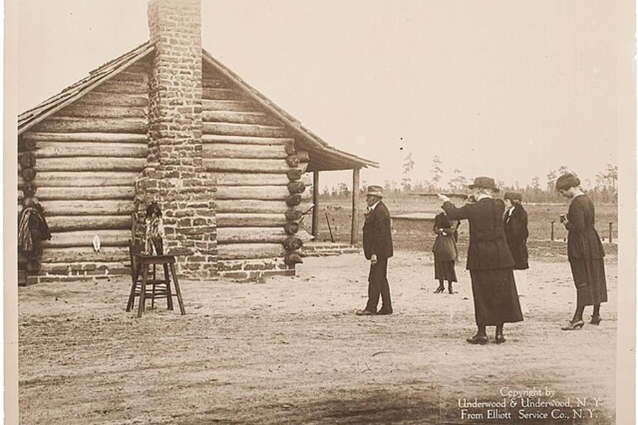 Sepia image of a woman aiming a pistol in an outdoor shooting practice session beside a rustic log cabin. A small dog stands on a stool in front of a chimney as a crowd of men and women watch from a distance, possibly observing a trick shooting demonstration.