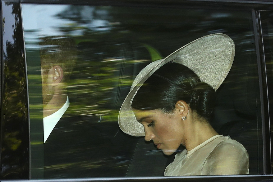 A woman in a light-colored outfit and wide-brimmed hat sits in the backseat of a car, looking down solemnly, with a man beside her barely visible through the tinted window. The reflection of trees and motion blur suggest the car is in transit.