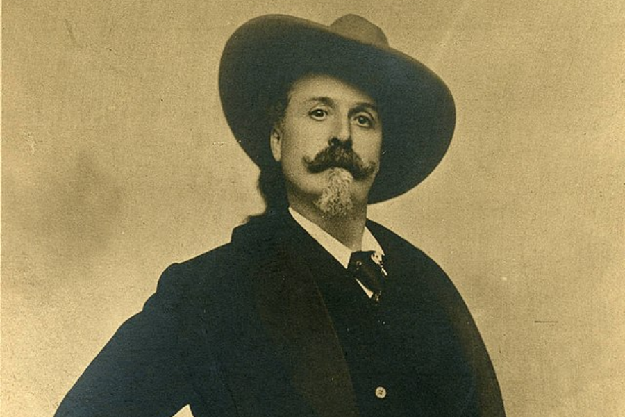 Sepia portrait of a distinguished man with a curled mustache and goatee, wearing a wide-brimmed cowboy hat and a dark suit with a vest and cravat. He stands confidently with one hand on his hip, exuding a classic Old West showman presence.