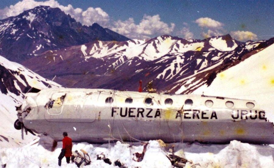A broken fuselage of a plane marked "FUERZA AEREA URUG" lies in the snow-covered Andes mountains. One person in a red jacket stands beside the wreckage, surrounded by white snow and rugged peaks under a clear blue sky.