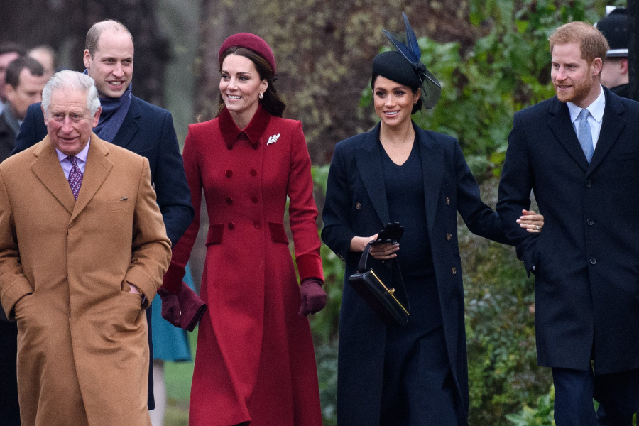 A group of royals walk together outdoors during a formal winter event. From left to right: a man in a camel coat and purple tie, a man in a navy coat smiling, a woman in a deep red coat and matching headband, a woman in a black coat and fascinator holding a clutch, and a man in a dark coat with a light blue tie walking arm in arm with her.