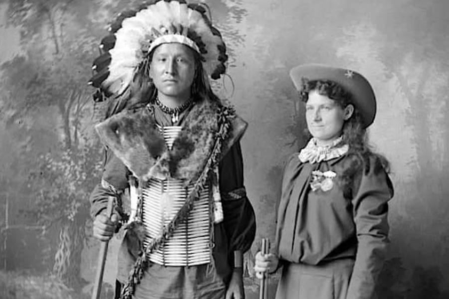 Black and white studio portrait of a Native American man in a traditional headdress and regalia standing beside a woman in Western-style clothing and a wide-brimmed hat, holding a rifle. The pair pose solemnly against a painted woodland backdrop, suggesting a promotional image or staged alliance.