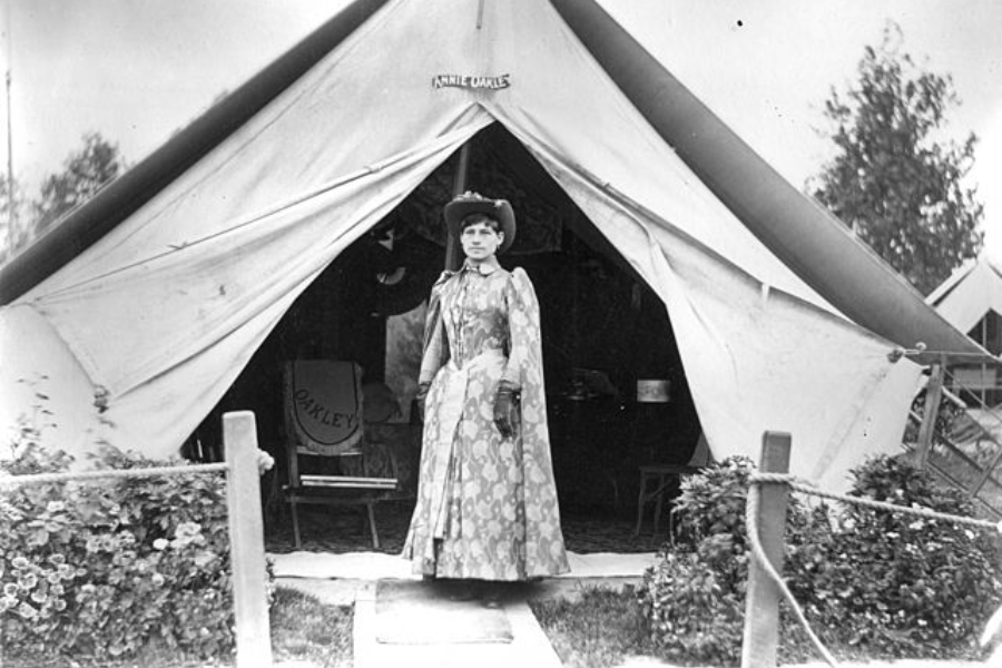 Black and white photograph of a woman standing in front of a large canvas tent labeled “Annie Oakley.” She wears a patterned full-length dress, gloves, and a Western-style hat, standing confidently on a small pathway flanked by ropes and flower bushes.