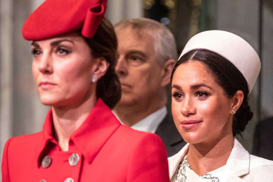 Two women stand side by side at a formal event, both dressed in elegant outfits with matching hats. The woman on the left wears a bright red coat with large silver buttons and a red pillbox hat, looking ahead seriously. The woman on the right wears a white coat with a structured white hat and a soft expression.