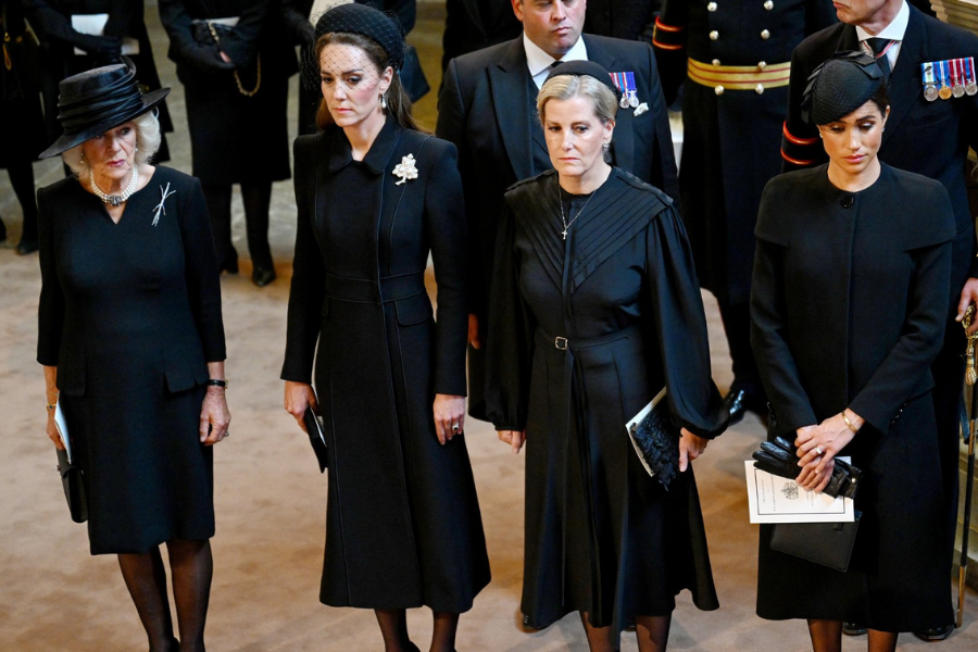 Four women dressed in black stand solemnly during a formal ceremony, likely a memorial or funeral. From left to right: a woman in a black dress and wide-brimmed hat, a woman in a long coat with a netted fascinator and floral brooch, a woman in a pleated black dress with a cross necklace, and a woman in a structured coat and black fascinator holding gloves and an order of service.