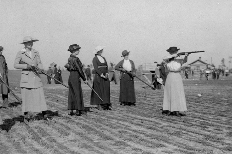 Black and white photograph of a line of women dressed in early 20th-century attire, participating in a shooting competition or practice. One woman is actively aiming and firing a shotgun, while the others stand ready with firearms on a plowed field, as spectators and a building can be seen in the background.