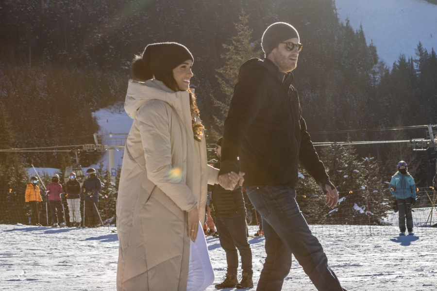 A couple holds hands while walking on a snowy slope at a ski resort, dressed warmly in winter clothing. The woman wears a long beige puffer coat and a black knit hat, while the man wears a black jacket, jeans, and a gray beanie, with ski lifts and other people in the background.