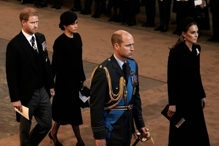 Four individuals walk in a formal procession during a solemn ceremonial event. The men are dressed in formal attire, one in a decorated military uniform and the other in a black morning suit with medals. The women wear black coats and hats, one with a veil and brooch, and the other carrying gloves and a program.