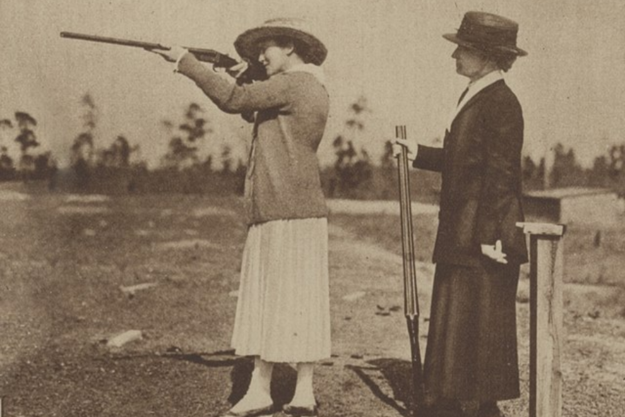 Sepia-toned photo of two women at an outdoor shooting range. One woman, dressed in a light-colored skirt and cardigan, is aiming a rifle while the other, in darker formal attire, stands beside her holding a second rifle.