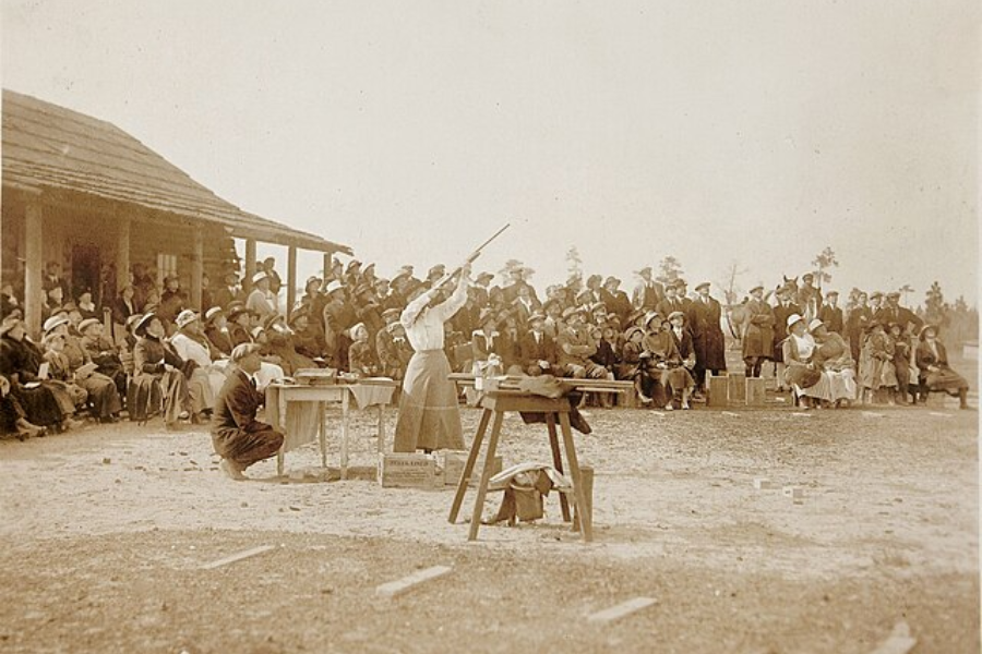 Sepia photograph showing a woman aiming a rifle skyward in front of a large outdoor audience, likely at a marksmanship or shooting exhibition. The crowd, dressed in early 20th-century attire, watches intently as the event unfolds near a rustic wooden building.