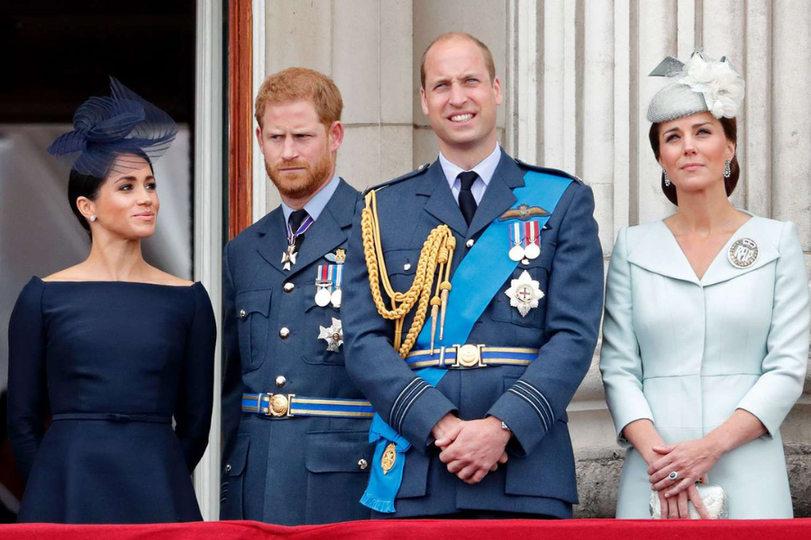 Four formally dressed individuals stand on a palace balcony during a ceremonial event. From left to right: a woman in a navy dress and fascinator, a man in a decorated Royal Air Force uniform, another man in a similar blue uniform with gold detailing, and a woman in a pale mint dress with a white floral hat and brooch.