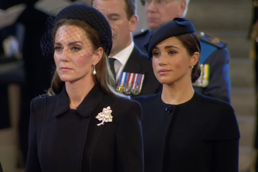 Two women dressed in black stand solemnly during a ceremonial event. The woman in front wears a dark outfit with a large jeweled brooch, pearl earrings, and a netted headpiece, while the woman behind her wears a simple black coat and a black fascinator with a small veil. Military personnel with medals are visible in the background.