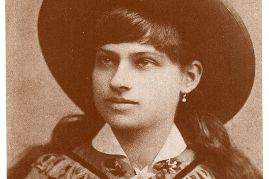Sepia-toned headshot of a young woman wearing a wide-brimmed Western hat and fringed clothing with a decorative bow. She has a calm, composed expression, a single drop earring, and neatly styled bangs.