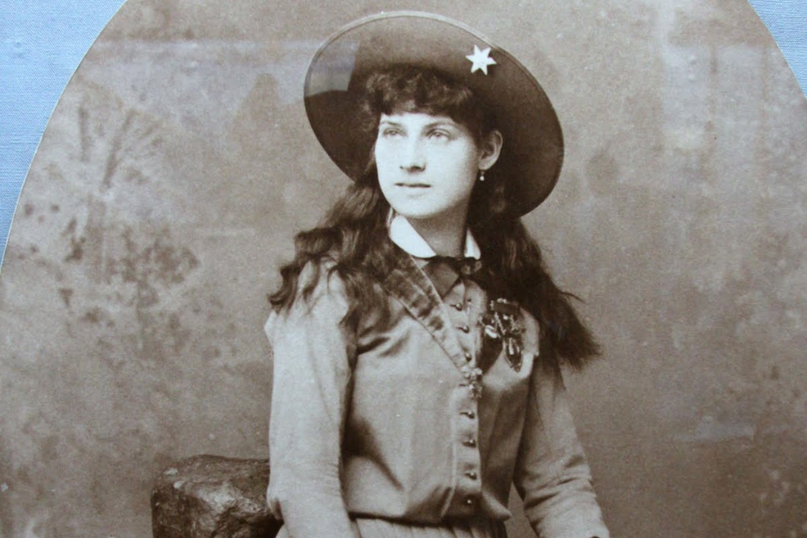 Sepia studio portrait of a young woman seated beside a rock prop, dressed in Western attire with a star-adorned hat and long curled hair. She wears a decorated blouse with medals and ribbons, gazing slightly off-camera with a composed expression.
