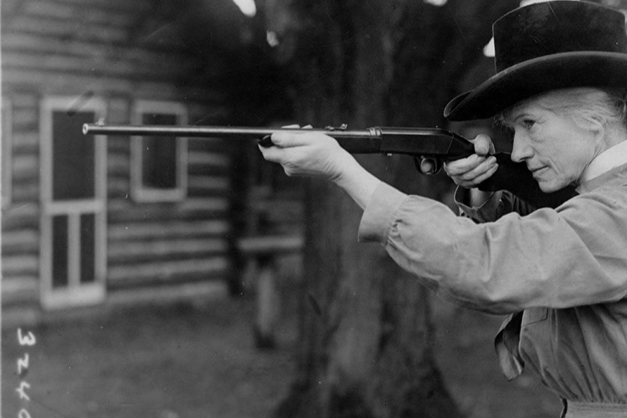 Black and white photograph of an older woman in a wide-brimmed hat and buttoned shirt, aiming a rifle with intense focus. She stands outdoors near a log cabin, framed by large trees in the background.