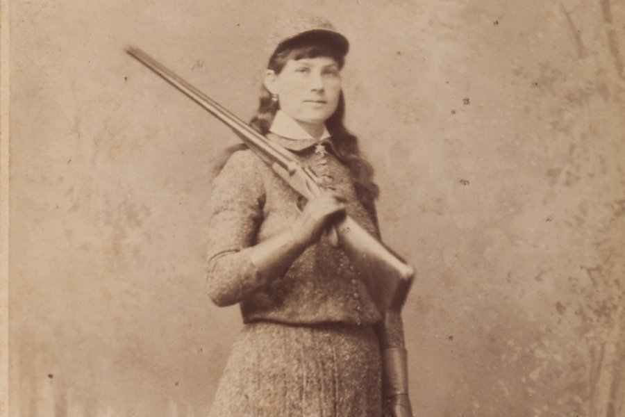 Sepia portrait of a young woman standing with a long rifle resting on her shoulder, dressed in a matching textured outfit with gloves and a flat-brimmed cap. She gazes confidently at the camera, set against a plain studio backdrop.