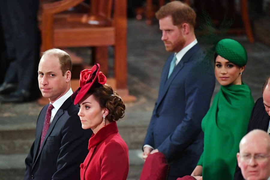 Four individuals dressed in formal attire attend a ceremonial event inside a grand venue. In the foreground, a man in a dark suit and a woman in a red coat with a dramatic floral hat sit side by side. Behind them, another man in a blue suit and teal tie stands next to a woman in a vibrant green cape and matching fascinator, looking ahead.