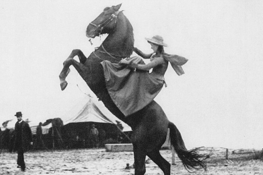 Black and white action photo of a woman in a flowing dress and wide-brimmed hat riding a horse that is rearing dramatically on its hind legs. A circus-style tent and onlookers are visible in the background, suggesting a Wild West performance or exhibition.