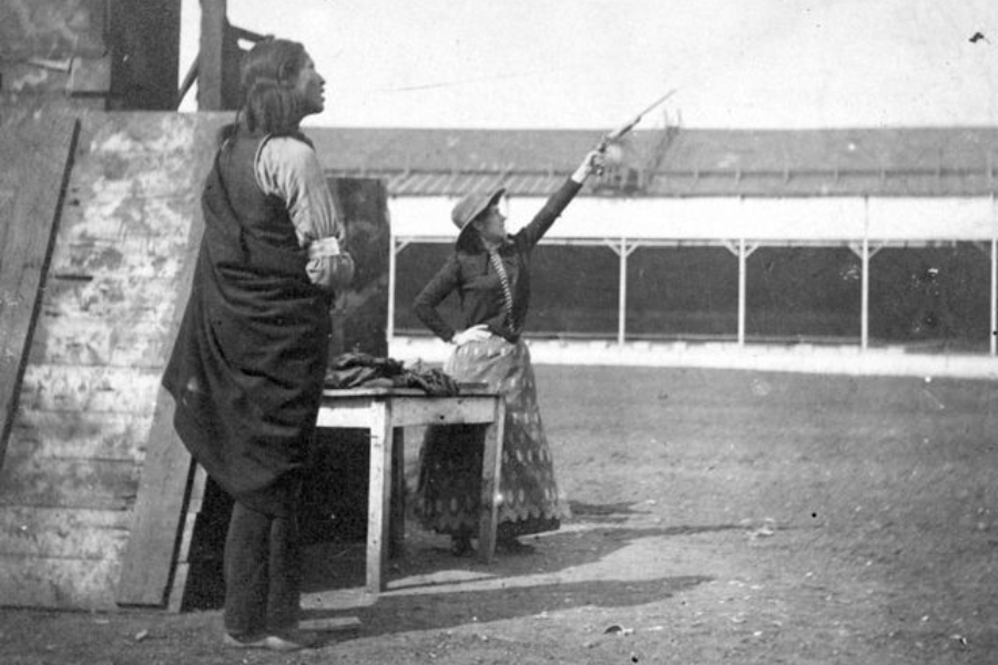 Black and white photograph of a woman in a patterned skirt and Western hat raising a pistol dramatically toward the sky, with a man in traditional Native American clothing standing nearby. They are positioned beside a rustic wooden platform, likely part of a Wild West show arena.