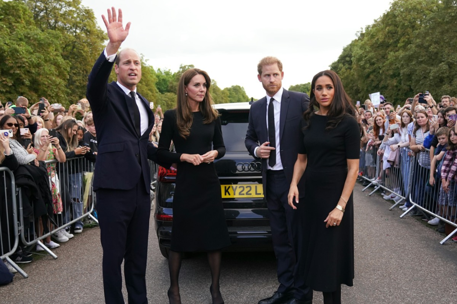 Two couples dressed in black stand in front of a dark car on a road lined with crowds and barricades. One man waves to the gathered public, while the others stand solemnly, acknowledging the crowd. The mood appears reflective, with all four individuals wearing formal mourning attire.