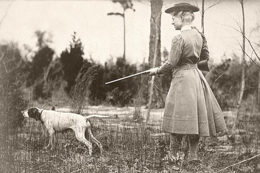 Sepia photograph of a woman dressed in a long, belted hunting coat and hat, holding a rifle as she walks through tall grass behind a pointing dog in a wooded area. The dog appears alert, possibly having caught a scent, as the woman follows attentively.