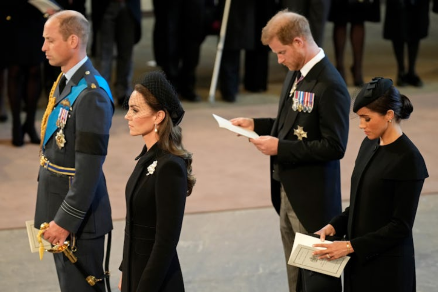 Four royals stand in solemn formation during a ceremonial event. The man in front wears a formal military uniform with medals and a ceremonial sword. Behind him, a woman in a black coat and netted fascinator stands with a serious expression. Two more individuals follow, reading from printed programs—one in a morning suit with medals and the other in a long black coat and structured hat.