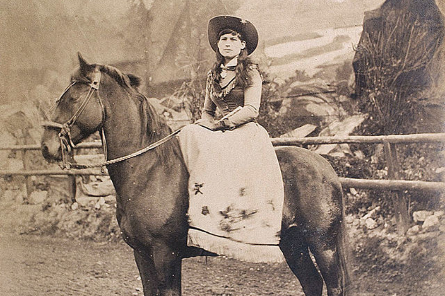 Vintage photograph of a poised woman in a cowboy hat and period Western attire sitting sidesaddle on a dark horse. She poses confidently in an outdoor setting enclosed by a wooden fence, suggesting a frontier or show environment.