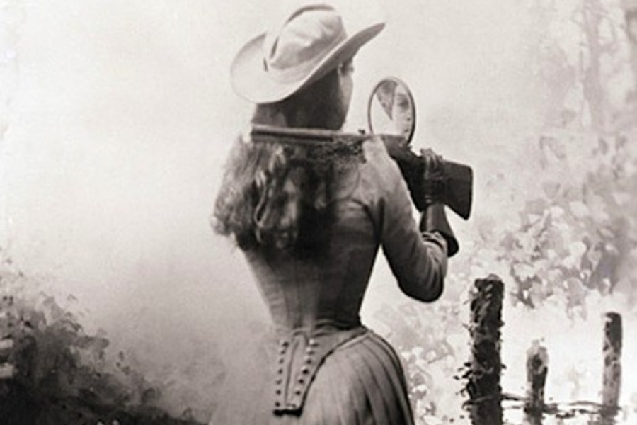 Vintage photograph showing a woman in a fitted dress and wide-brimmed hat holding a rifle over her shoulder, aiming using a small round mirror to shoot backward. Her reflection is visible in the mirror, capturing the precision and theatrical flair of her sharpshooting trick.