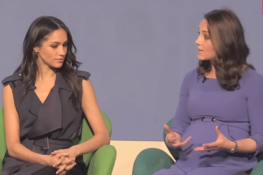 Two women are seated onstage in modern, colorful chairs during a formal discussion. The woman on the left wears a sleeveless navy dress, listening intently with her hands folded. The woman on the right, visibly pregnant in a fitted lavender dress, gestures with both hands as she speaks.