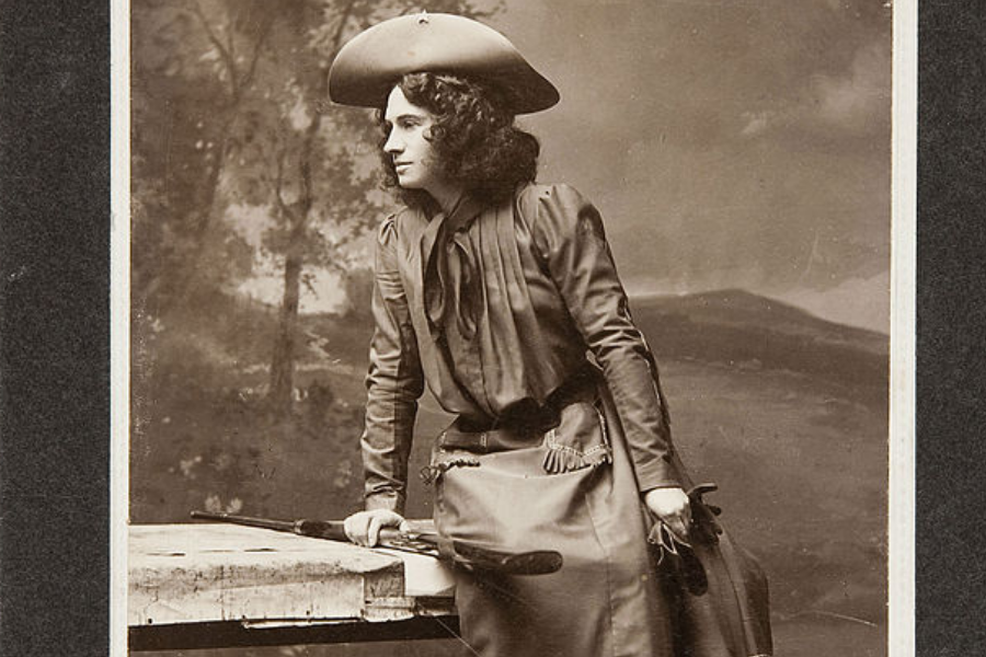 Sepia studio portrait of a woman in a wide-brimmed Western hat and fringed outfit, leaning casually on a wooden table with a rifle resting beside her. She gazes thoughtfully to the side against a painted backdrop of trees and rolling hills.
