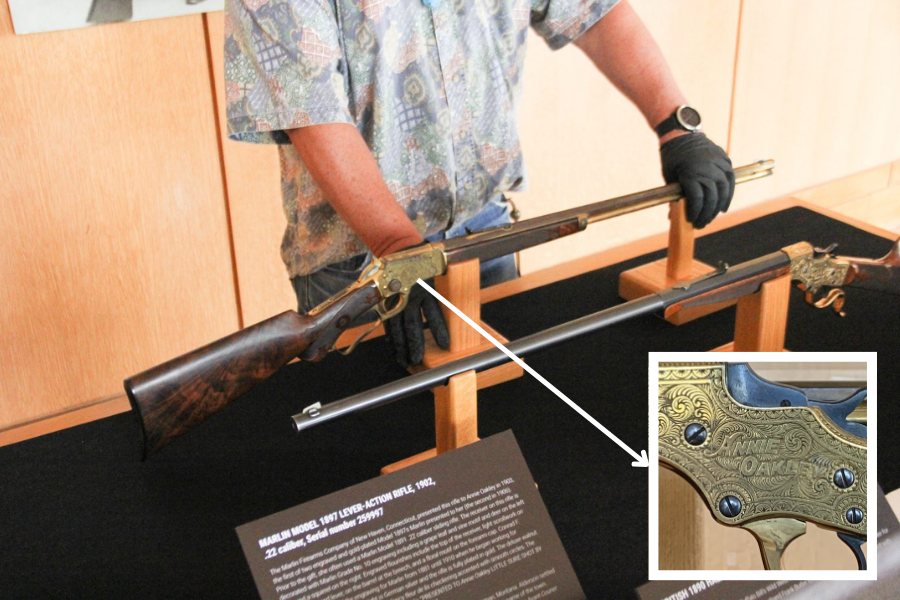 A museum staff member wearing gloves handles an ornately engraved Marlin Model 1897 lever-action rifle displayed on a wooden stand. The zoomed-in inset reveals intricate gold detailing with the engraved name “Annie Oakley,” identifying it as her personal firearm from 1902.