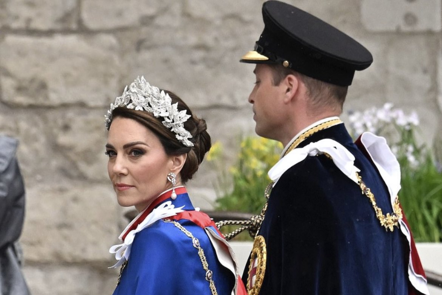 A woman wearing a dazzling silver tiara, drop earrings, and a royal blue ceremonial robe looks over her shoulder with a poised expression. Beside her, a man in a dark uniform with gold trim and a peaked cap stands in profile, both attending a formal royal event outside a stone building.