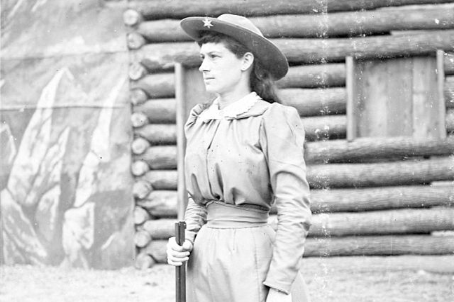 Black and white photograph of a woman in a high-collared dress and star-adorned hat standing in front of a log cabin. She holds a long rifle vertically by her side, gazing sternly into the distance with a composed and confident posture.