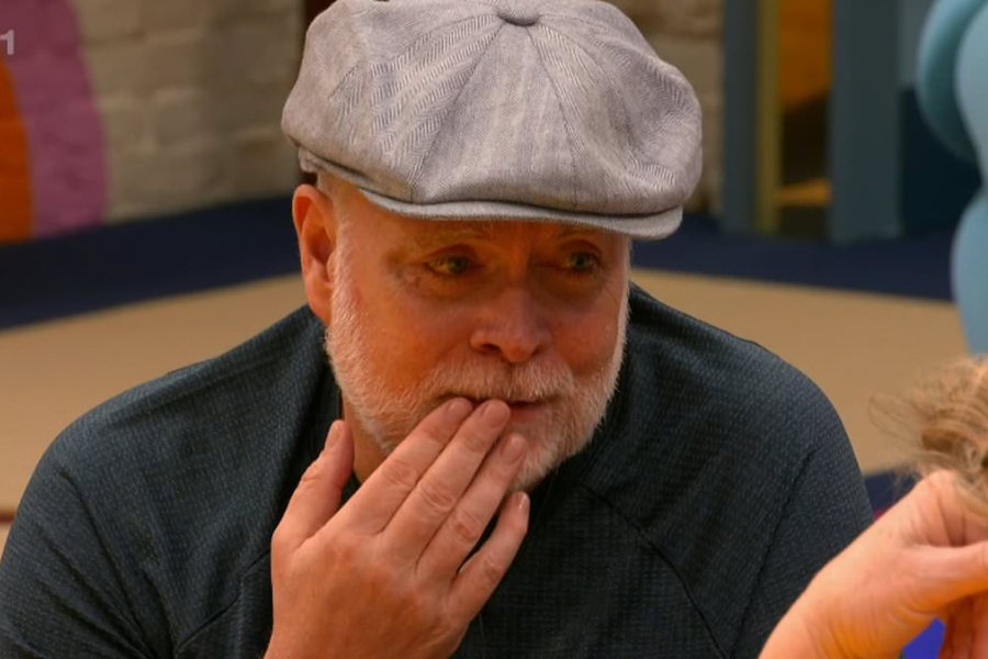 An older man with a gray beard and flat cap rests his hand gently against his face while speaking to someone out of frame. His expression is warm and slightly emotional, suggesting a heartfelt or reflective moment in a cozy indoor setting.