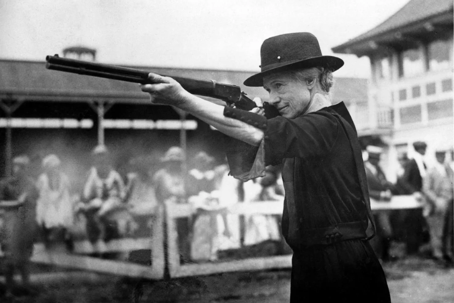 Black and white photograph of an elderly woman wearing a dark dress and hat, aiming a rifle with sharp concentration at an outdoor event. A blurred crowd of spectators watches from behind a fence, suggesting a public shooting demonstration or competition.