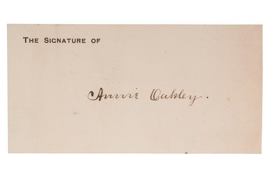 A cream-colored card with the printed text "THE SIGNATURE OF" at the top left, followed by a handwritten cursive signature reading “Annie Oakley” in brown ink. The autograph is centered neatly on the card.