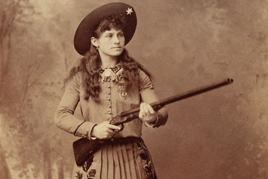 Sepia-toned studio portrait of a young woman with long curly hair, wearing a wide-brimmed hat with a star emblem and a pleated skirt. She confidently holds a rifle, dressed in embroidered Western-style clothing adorned with medals and decorative details.