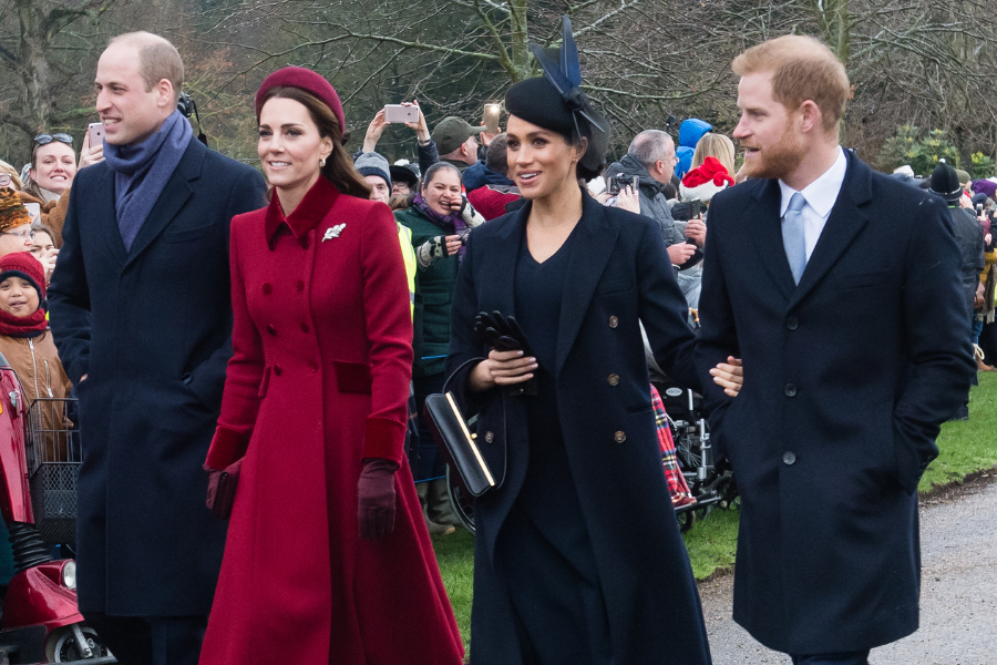 Four people walk together outdoors during a formal holiday event, surrounded by a crowd. From left to right: a man in a navy overcoat and scarf, a woman in a deep red coat and matching headband, a woman in a black coat with a fascinator and gloves, and a man in a dark overcoat with a light blue tie.