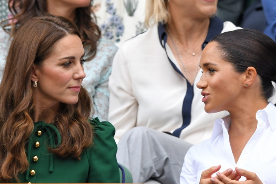 Two women sit next to each other in a crowded stadium, engaged in a conversation. The woman on the left wears a green blouse with gold buttons and has her hair down in waves, while the woman on the right wears a white collared shirt and gestures with her hands as she speaks.