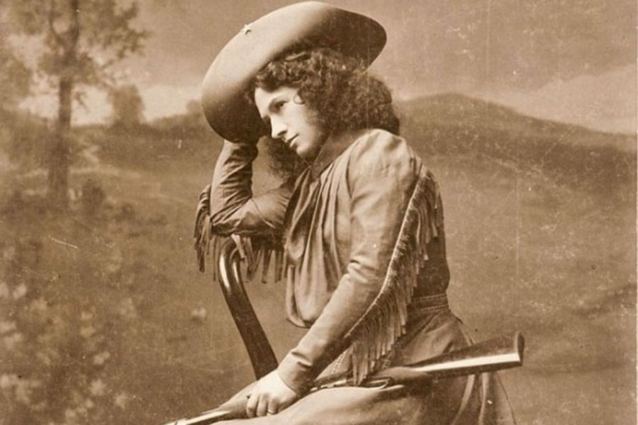 Sepia portrait of a contemplative woman seated backward on a wooden chair, resting her head on one arm. She wears fringed Western attire and a wide-brimmed hat, with a rifle resting across her lap against a painted countryside backdrop.