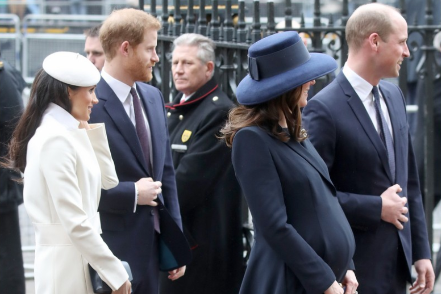 Two formally dressed couples stand outside in conversation near a gated entrance. On the left, a woman in a white coat and matching hat smiles beside a man in a navy suit; on the right, a visibly pregnant woman in a navy outfit and wide-brimmed hat chats with a man in a matching navy suit.