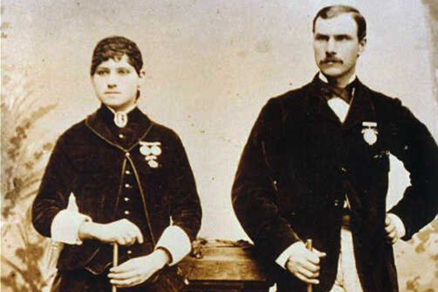 Sepia photograph of a woman and man dressed in formal 19th-century attire, both wearing medals pinned to their jackets. They each hold a cane or riding crop and pose side by side in a studio setting, suggesting a shared achievement or affiliation.