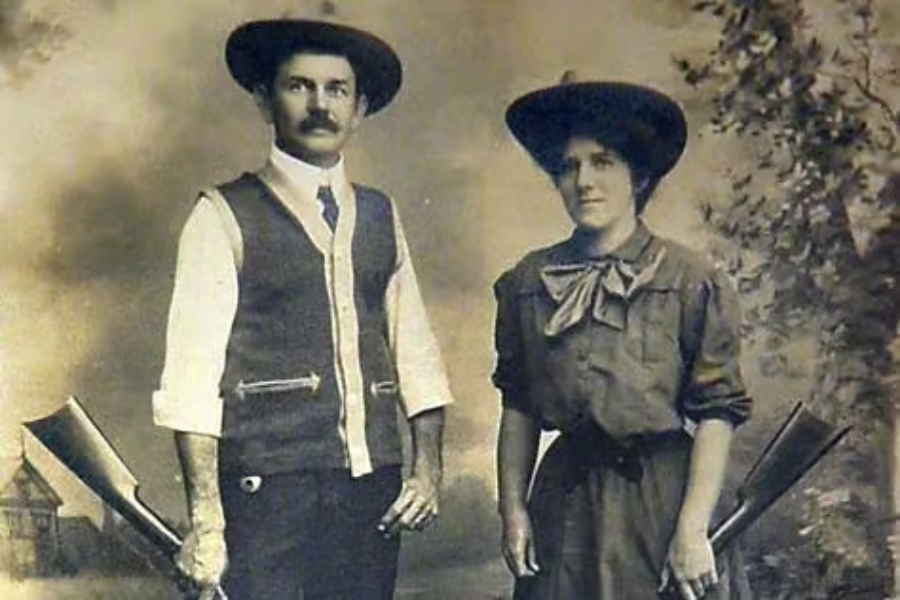 Faded vintage portrait of a man and woman standing side by side, each holding a shotgun. Both wear wide-brimmed hats and rugged attire, with the man in a vest and the woman in a dress with a large bow tie, suggesting they are hunters or sharpshooters.