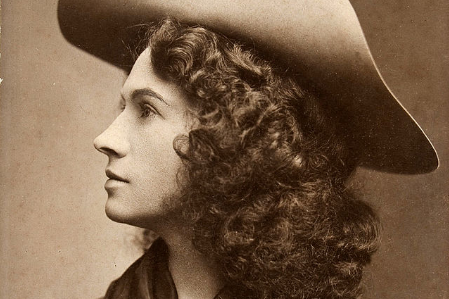 Sepia close-up profile portrait of a young woman with voluminous curly hair wearing a wide-brimmed Western-style hat. Her expression is calm and contemplative, with soft studio lighting highlighting her features.