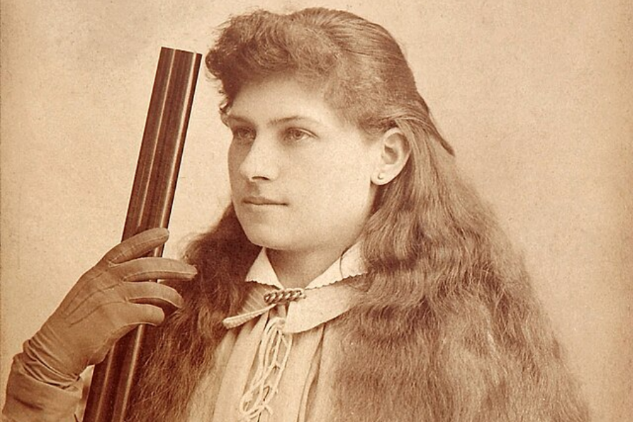 Sepia studio portrait of a young woman with long, wavy hair, wearing a lace-up blouse and a bow-tied collar. She holds a double-barrel shotgun upright beside her face with a gloved hand, looking off camera with a composed expression.