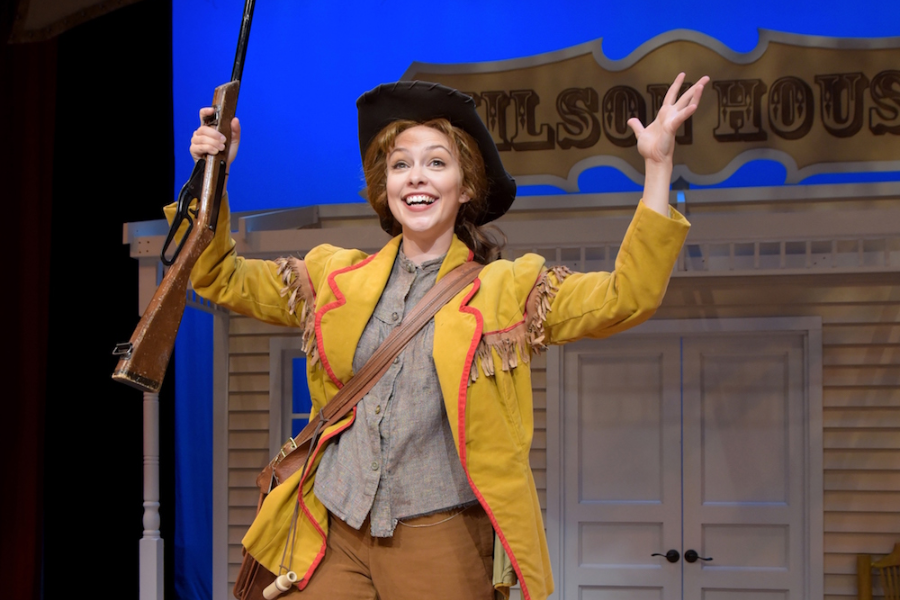 A smiling woman in a theatrical costume portraying Annie Oakley stands on stage holding a rifle aloft in one hand and gesturing joyfully with the other. She wears a yellow fringed jacket with red trim, brown pants, and a cowboy hat, with a Western building façade labeled "WILSON HOUSE" in the background.