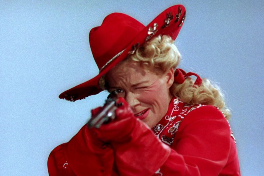 Vibrant color still of a woman in a bright red, rhinestone-embellished Western outfit and matching cowboy hat aiming a pistol while winking. Her blonde curls and theatrical pose suggest a Hollywood portrayal of Annie Oakley.